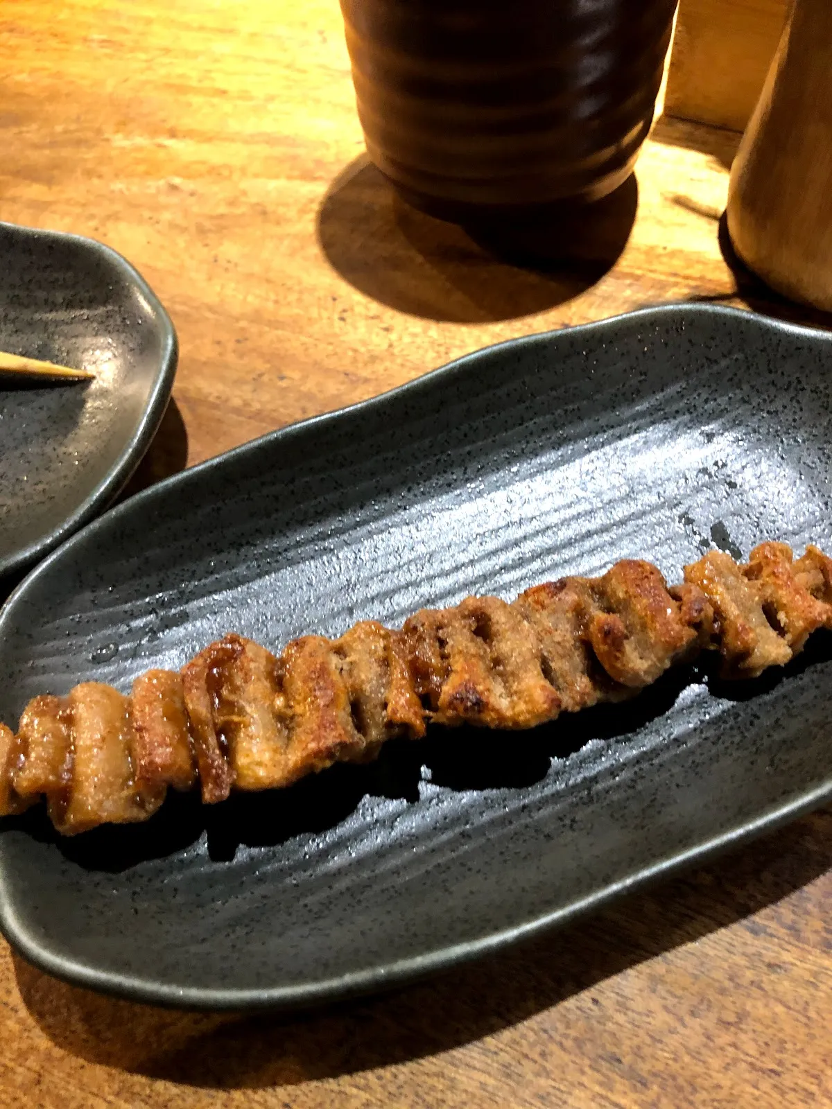 長條狀的烤豬五花串，薄肉片捲成波浪狀排滿整支竹籤，盛在深色長盤上