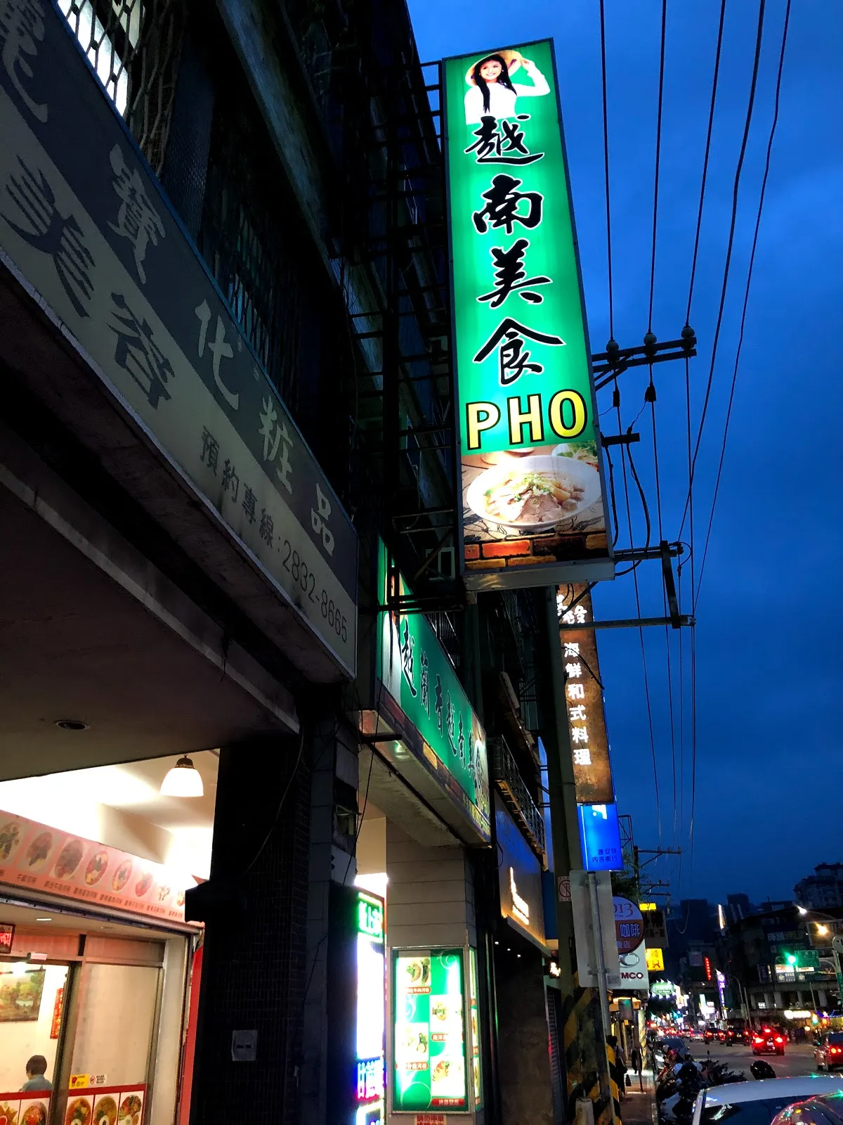 夜晚的街景，越南美食 PHO 的綠色直立招牌亮著燈，周圍是其他店家招牌與交錯的電線