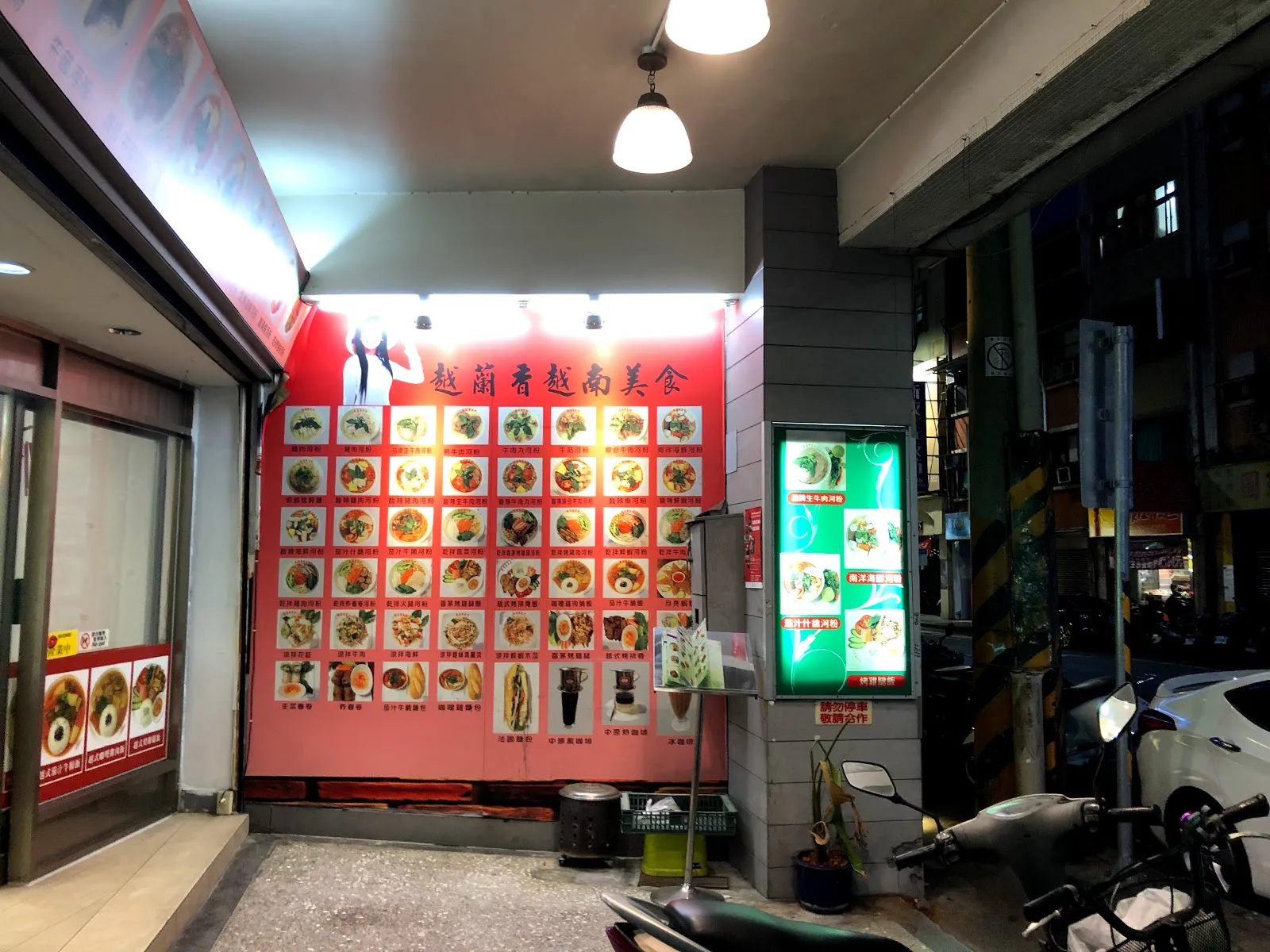 店門口的半戶外走道，粉紅色牆面貼滿餐點照片看板，右側是亮著燈的立式招牌
