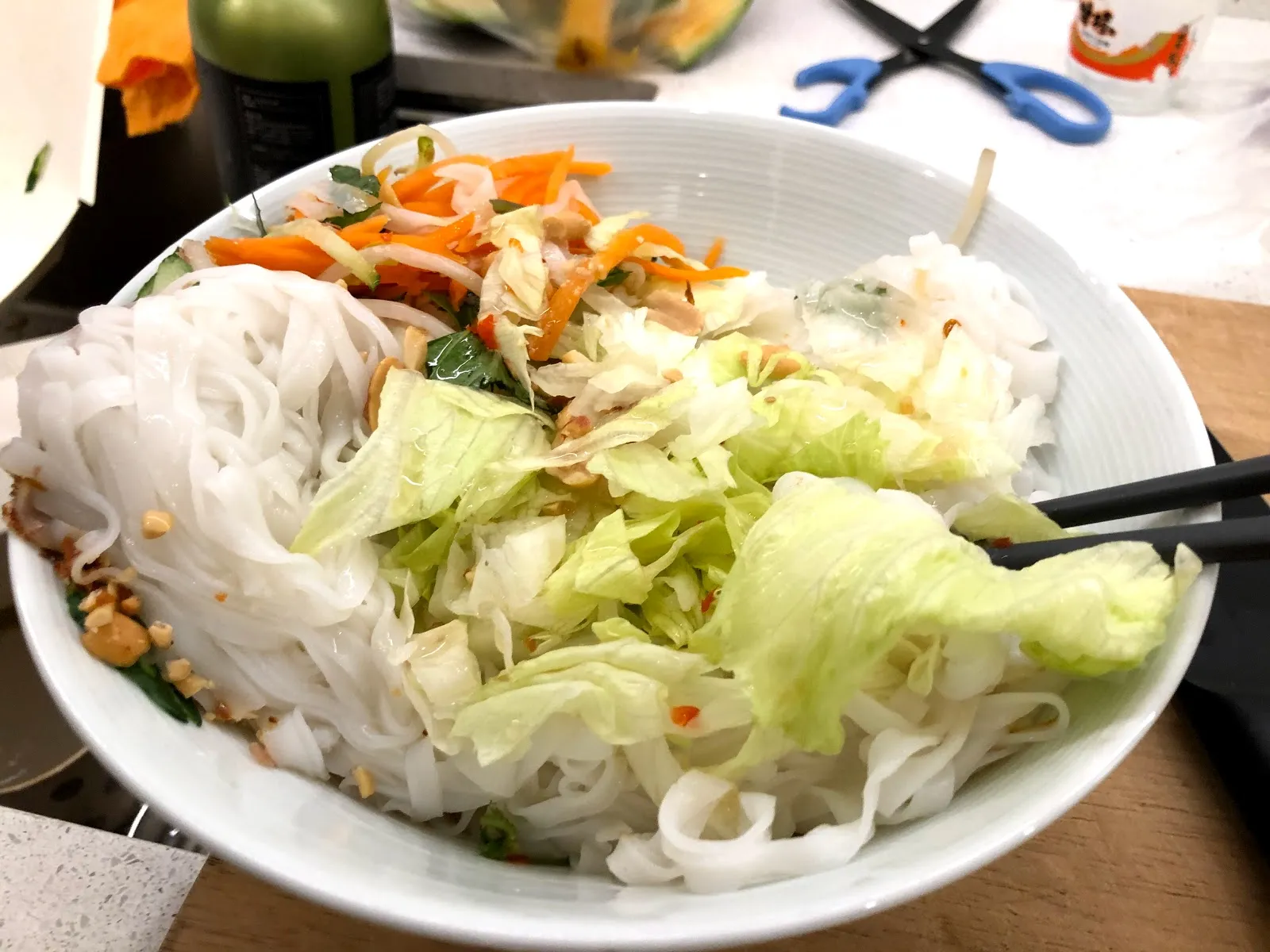 白色大碗裡的越南河粉，寬扁粉條上鋪滿生菜絲、紅蘿蔔絲與花生碎，插著一雙筷子