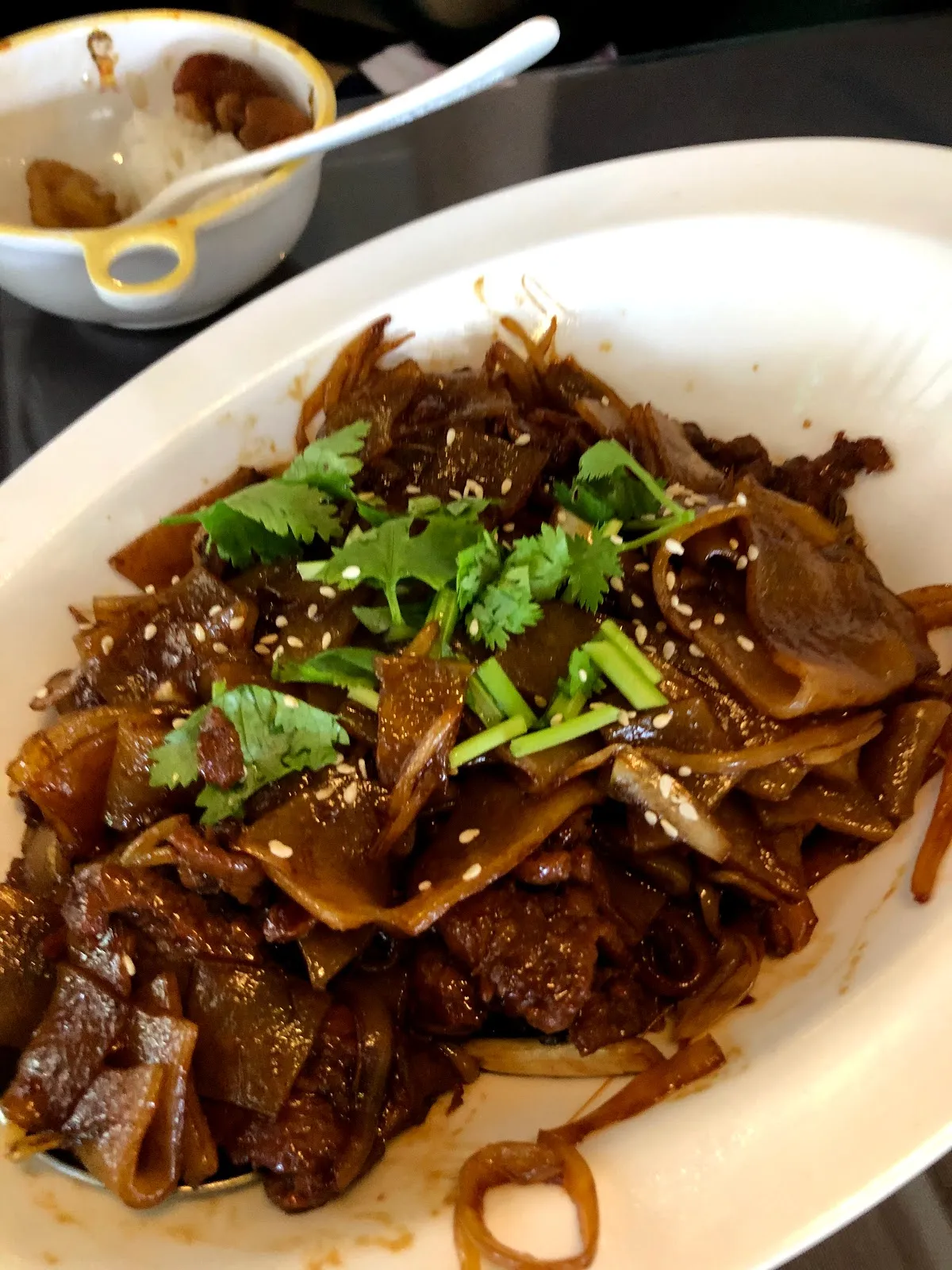 白盤上的港式乾炒牛河，寬版河粉與牛肉炒得油亮，撒上白芝麻與香菜，後方是一碗白飯