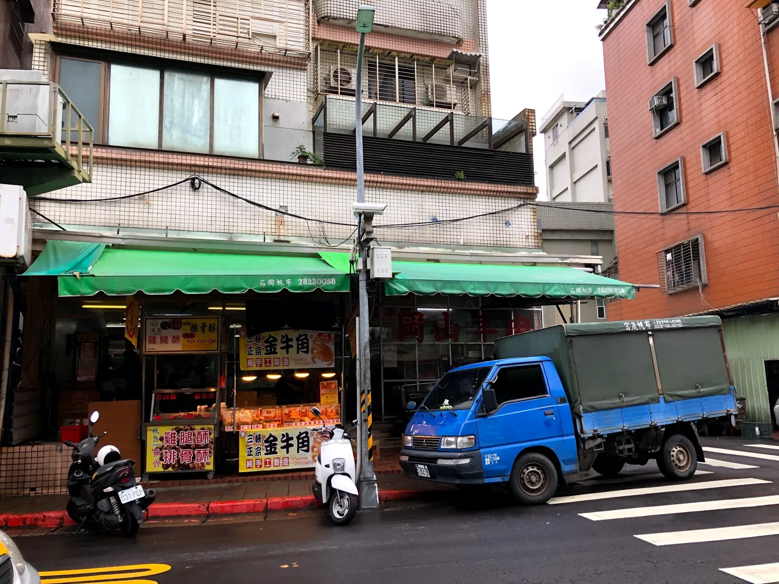 店面外觀，綠色遮陽棚下是紅底黃字的攤位招牌，門口停著兩輛機車，路邊停著一輛藍色小貨車
