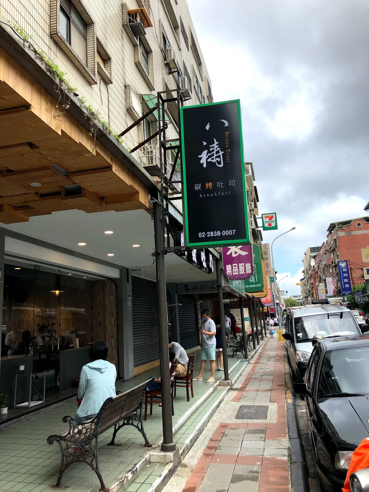 八禱炭烤吐司的店面外觀，綠底白字的直立招牌掛在騎樓柱上，人行道旁停著車，後方看得到 7-11 的招牌