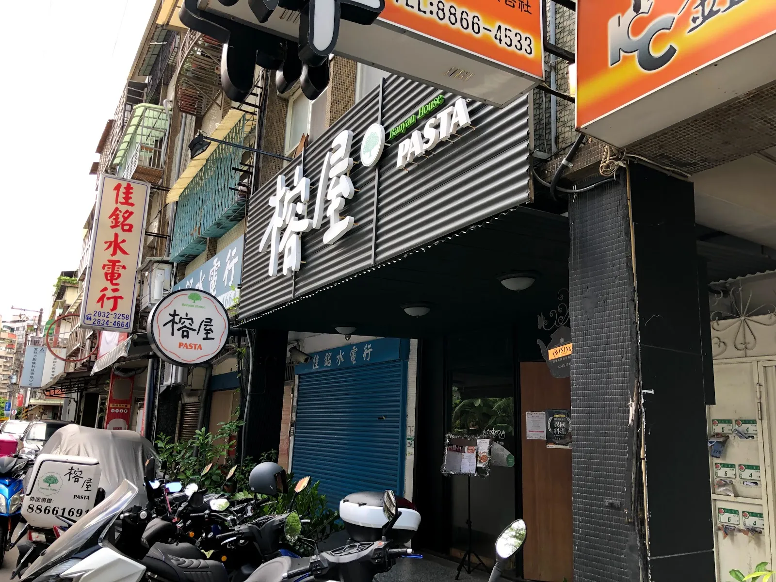 榕屋 PASTA 的店面外觀，黑色浪板招牌上是白色的「榕屋」二字，騎樓下停著幾輛機車，隔壁是水電行