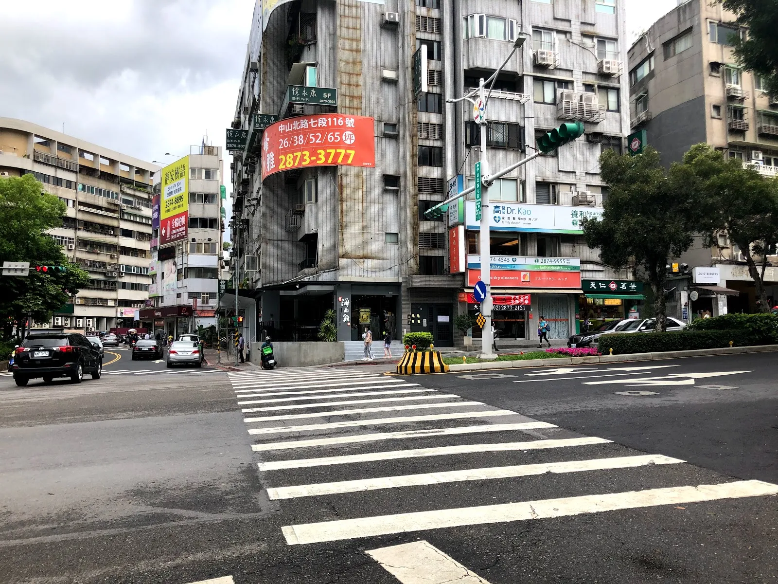 店外的十字路口街景，對面大樓外牆掛著紅色的建案廣告布條，路面畫著斑馬線，遠方有車輛等紅燈