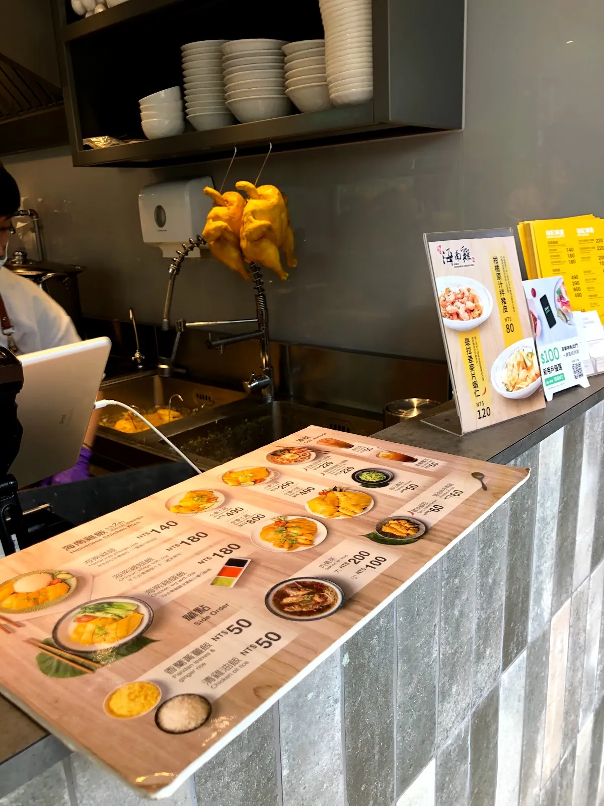店內點餐區，磁磚檯面上放著木紋餐點看板，後方掛著三隻黃澄澄的白斬雞，架上疊著白色碗盤