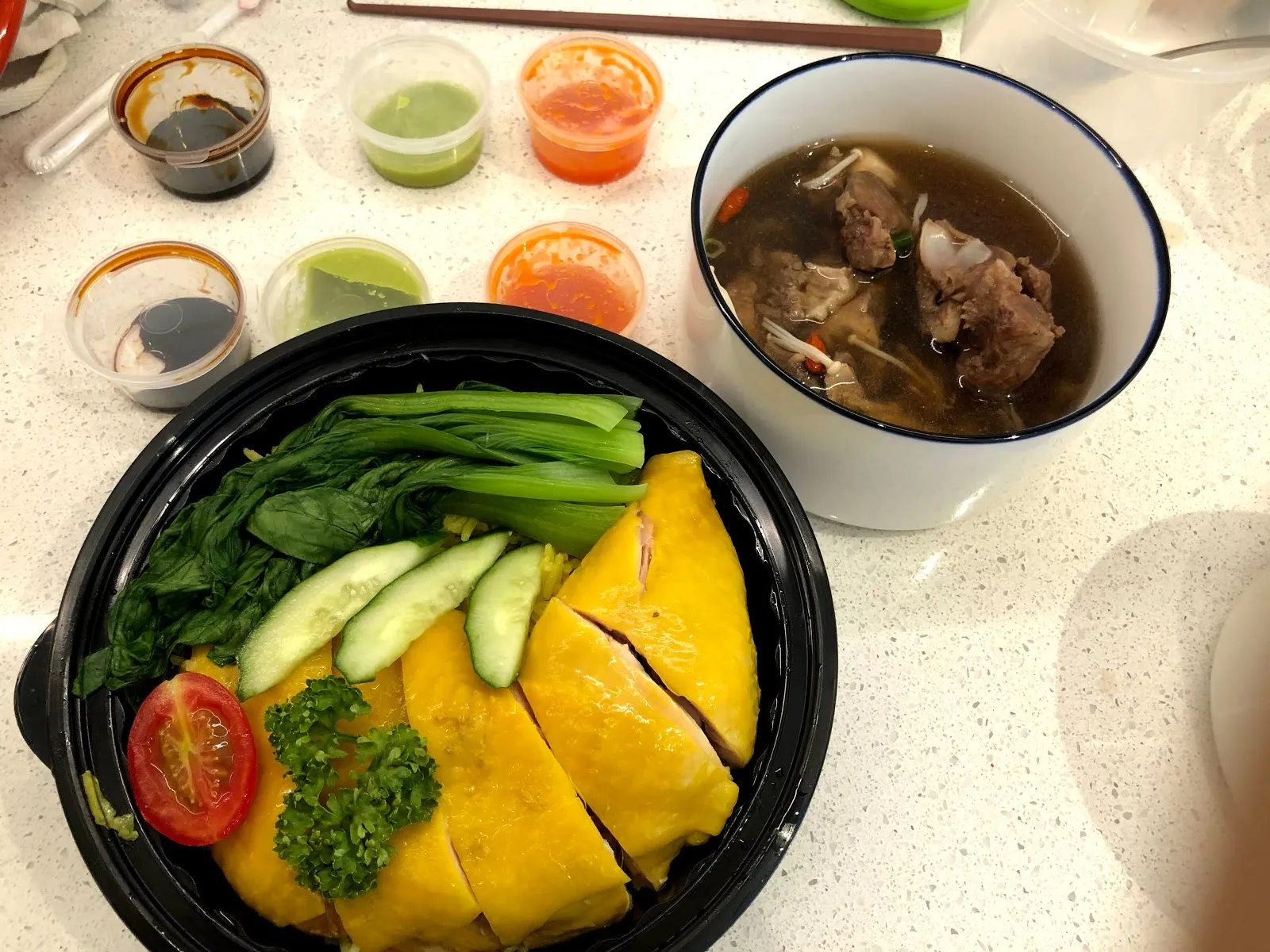 外帶回來的海南雞飯攤在桌上，黑色圓盒裡是雞肉、青江菜與小黃瓜，旁邊是一碗藥燉排骨湯與五杯不同顏色的沾醬