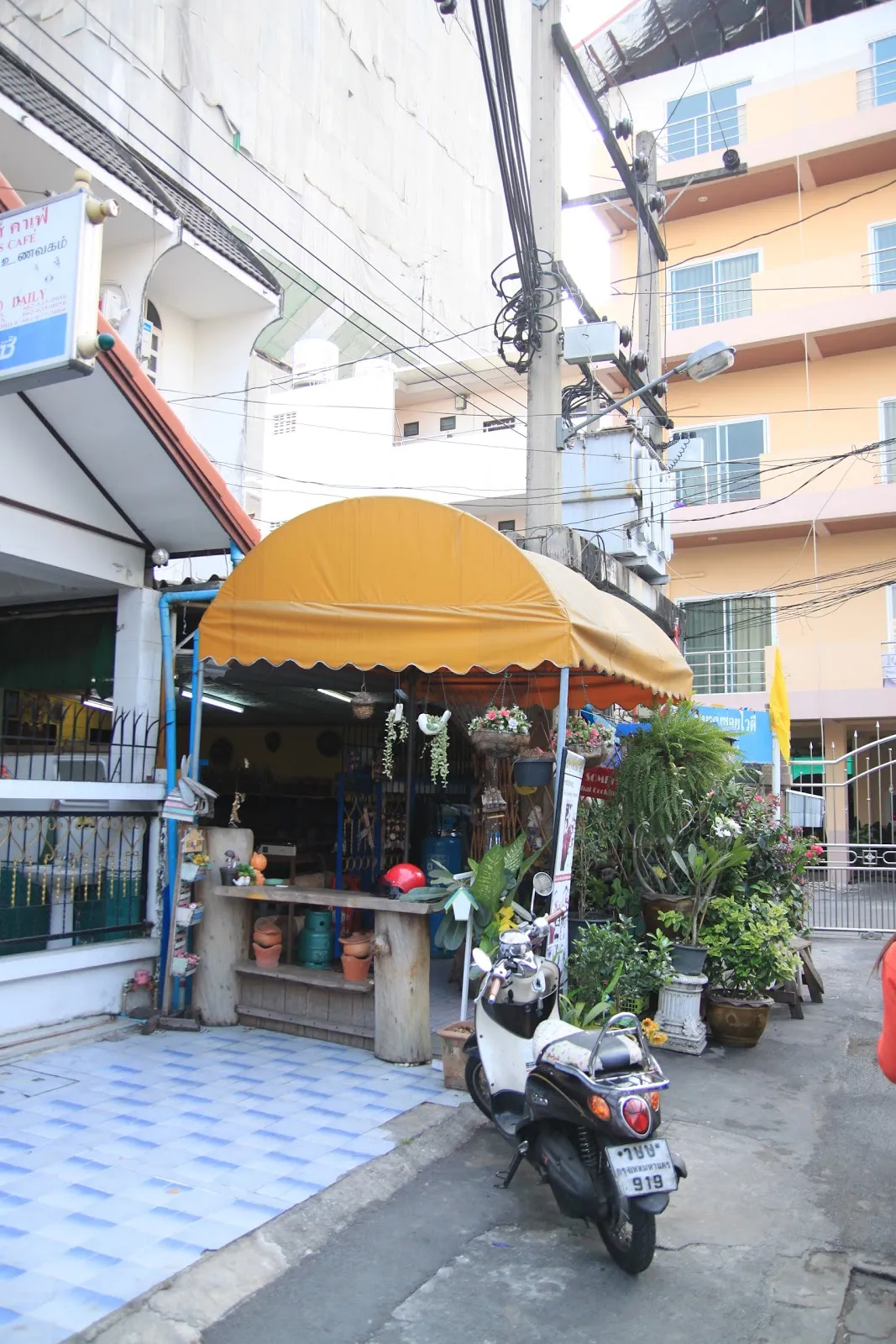街邊的店家，黃色遮陽棚下是磁磚地與盆栽，一輛機車停在門口，後方是黃色與粉色的樓房