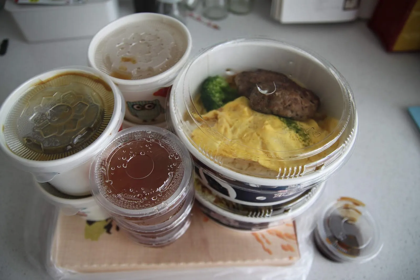 外帶回家的三份餐點攤在流理臺上,塑膠碗裡裝著漢堡排滑蛋飯,旁邊是清湯、咖哩醬與一小杯醬汁