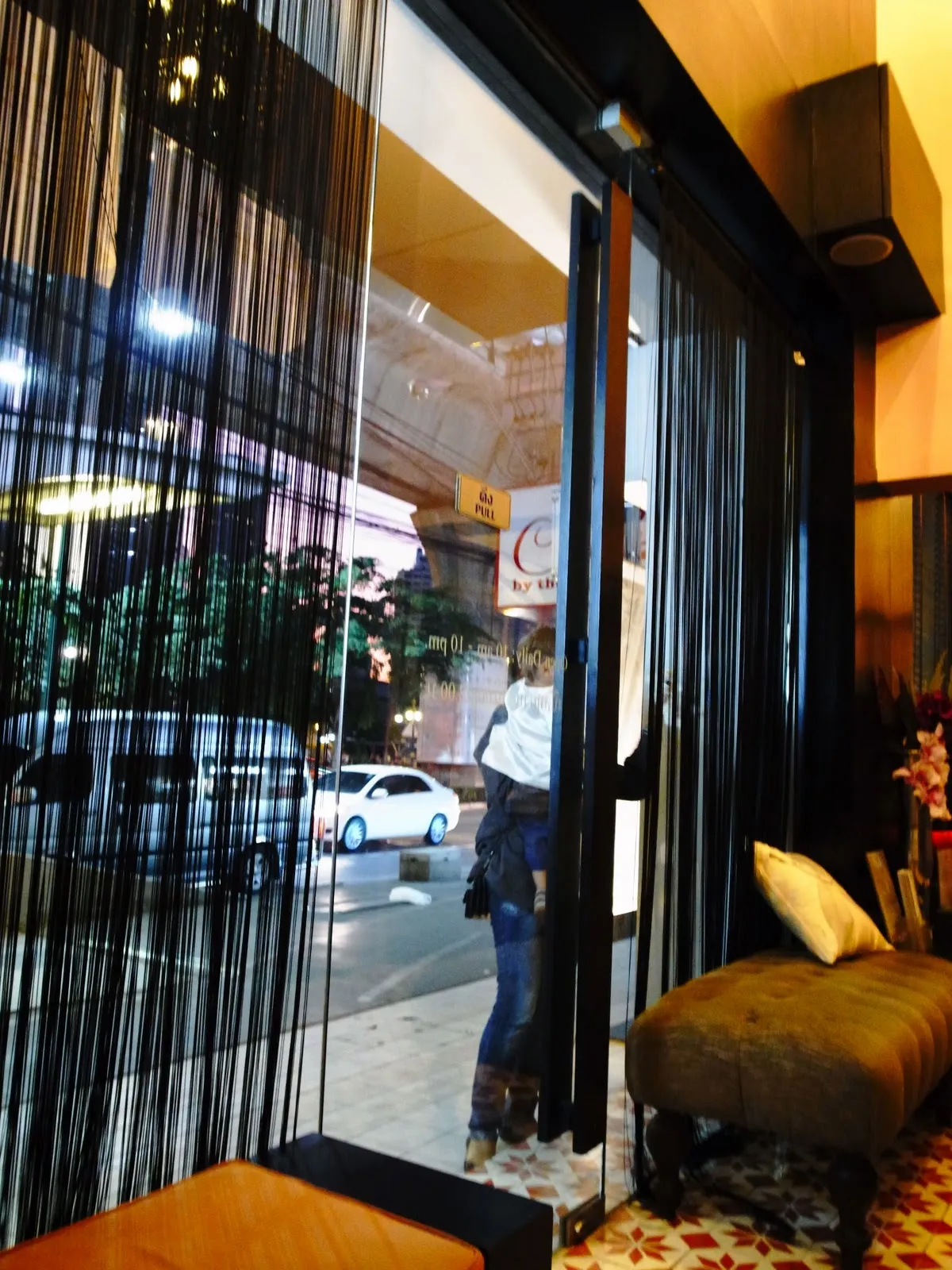 從店內往外看,垂掛的黑色珠簾旁是玻璃門,門外是街道與停著的白色汽車,室內擺著絨布長凳