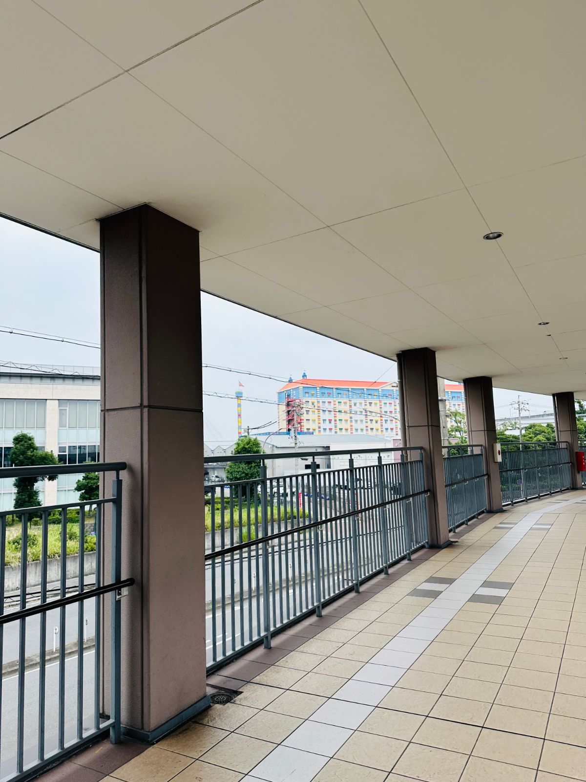 車站的半戶外連通道，一排方柱沿著欄杆延伸，遠方看得到樂高樂園色彩鮮豔的建築