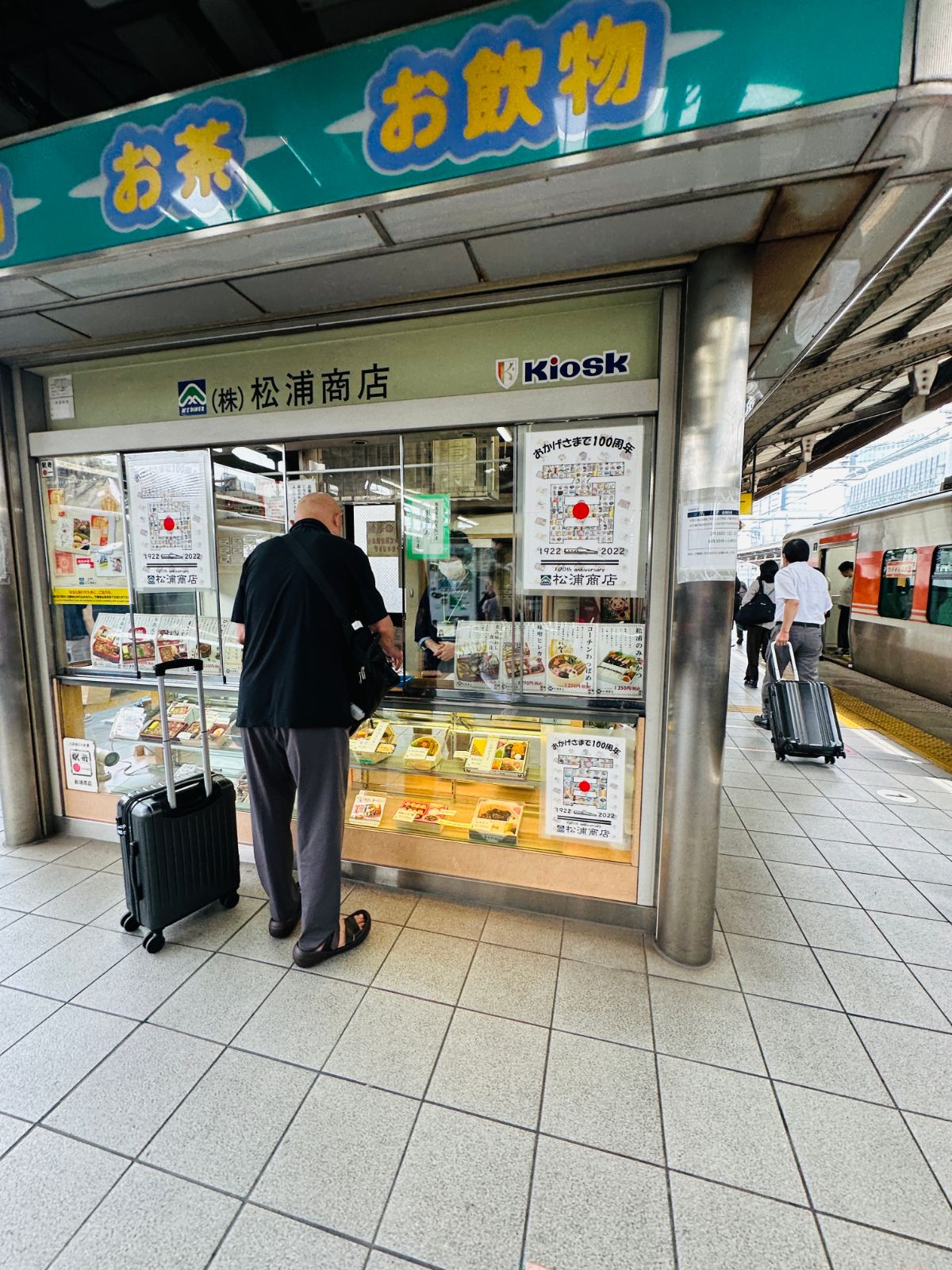 車站月台上的松浦商店 Kiosk，一名男子推著行李箱站在櫃檯前挑選便當，招牌上寫著「お茶」「お飲物」