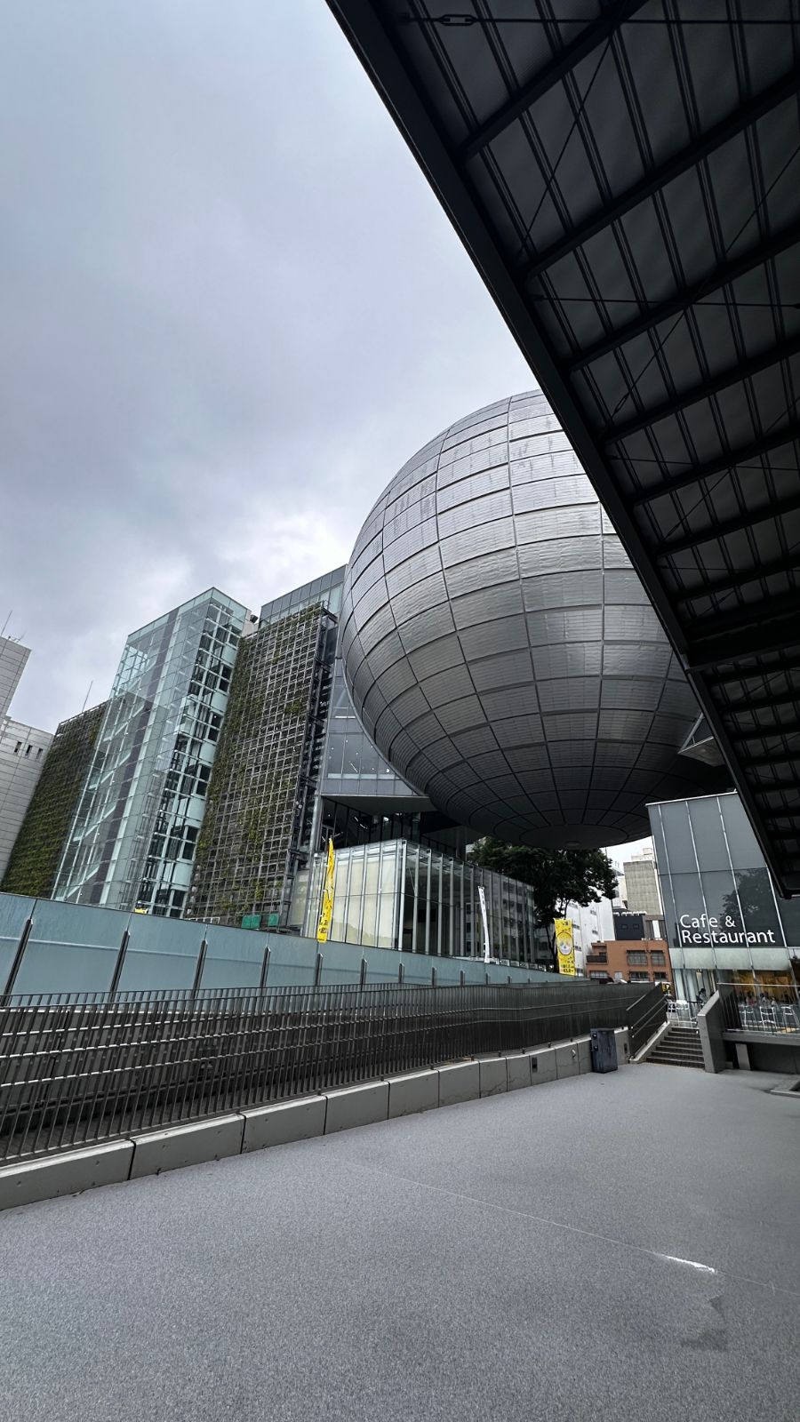 名古屋市科學館的外觀，巨大的銀色球體天文館嵌在建築中間，左側是覆蓋植生牆的大樓，天色陰沉