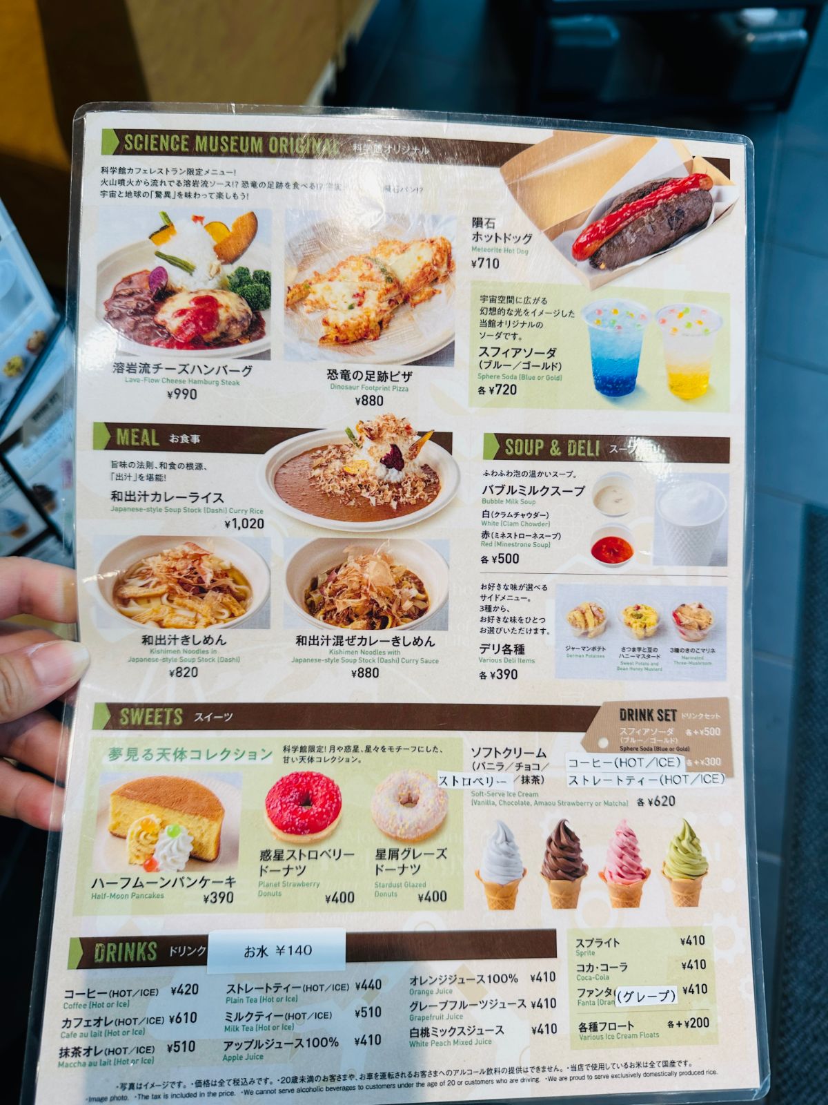 手拿著科學館餐廳的菜單，上面有隕石熱狗 710 圓、熔岩起司漢堡排 990 圓、恐龍足跡披薩 880 圓等餐點照片與價格