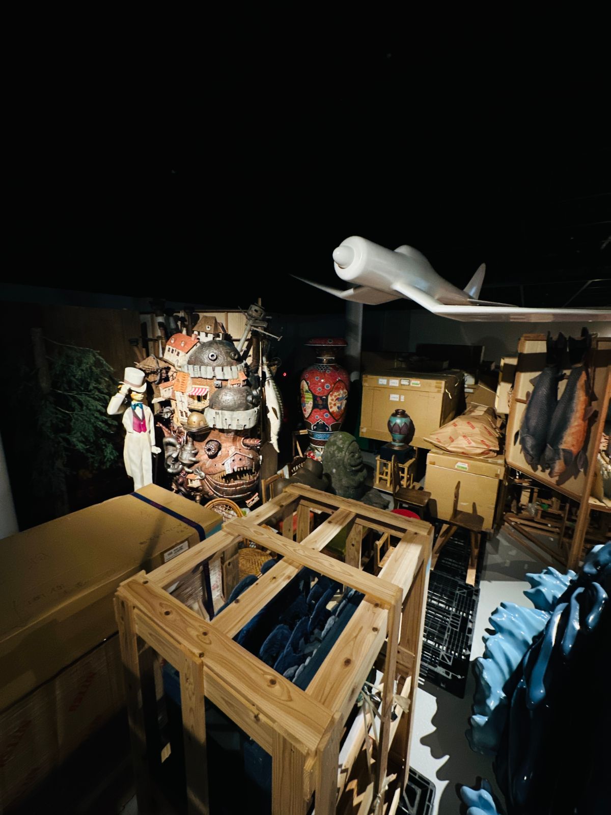 昏暗的倉庫展示區,堆滿木箱與道具,中央立著哈爾的移動城堡模型,上方懸著一架白色滑翔翼