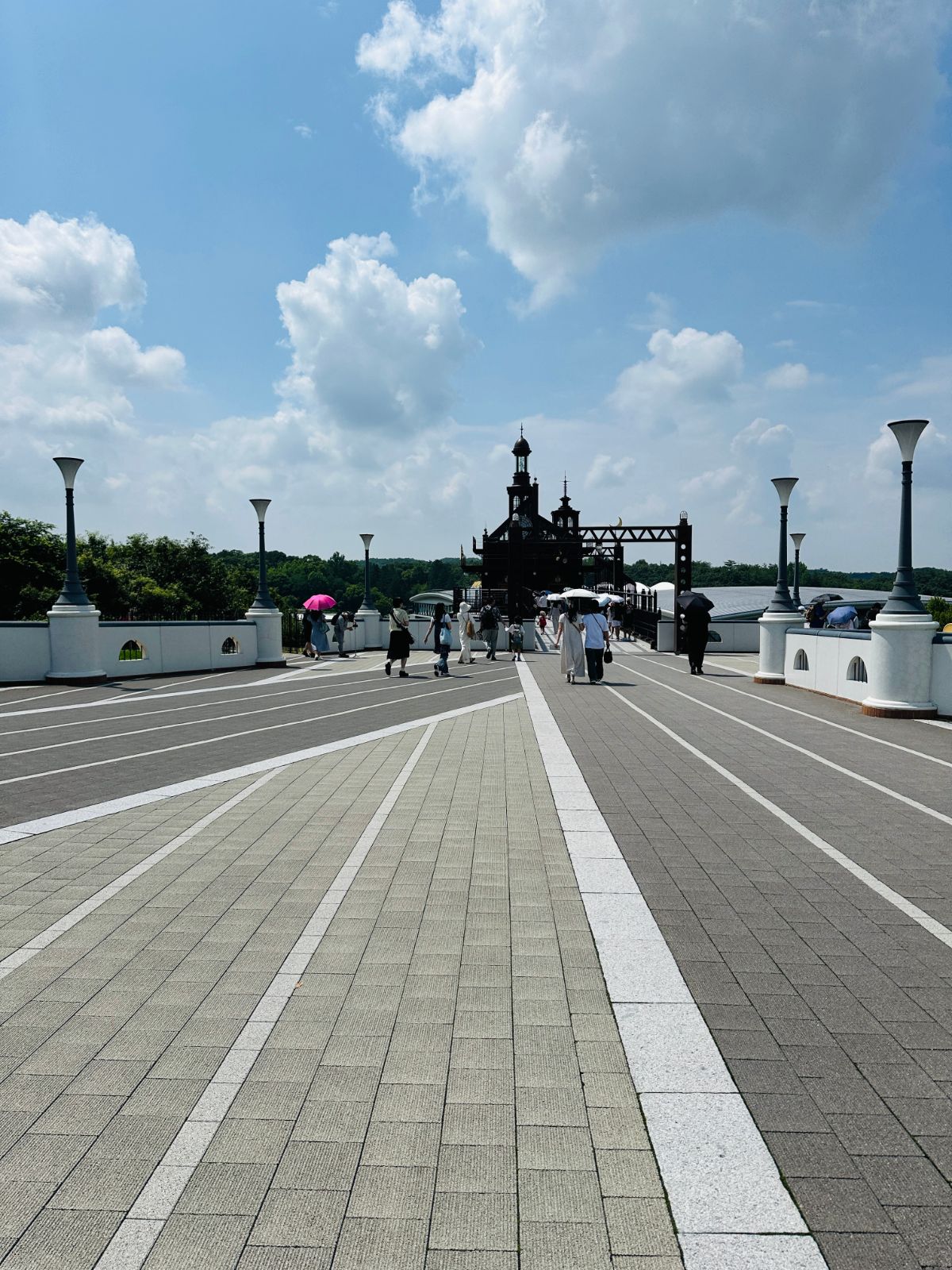 通往吉卜力公園的寬闊石板大道,兩側立著白色路燈,遠處是深色的鐘塔建築,天空有大片積雲