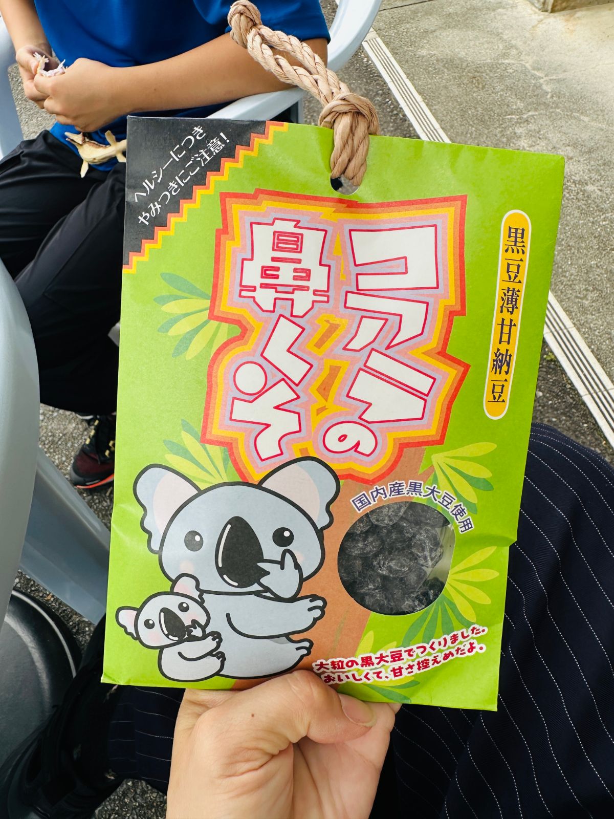 手上拿著一包無尾熊造型的黑豆甘納豆零食，綠色包裝上印著無尾熊母子插畫，開窗處看得到裡面的黑豆