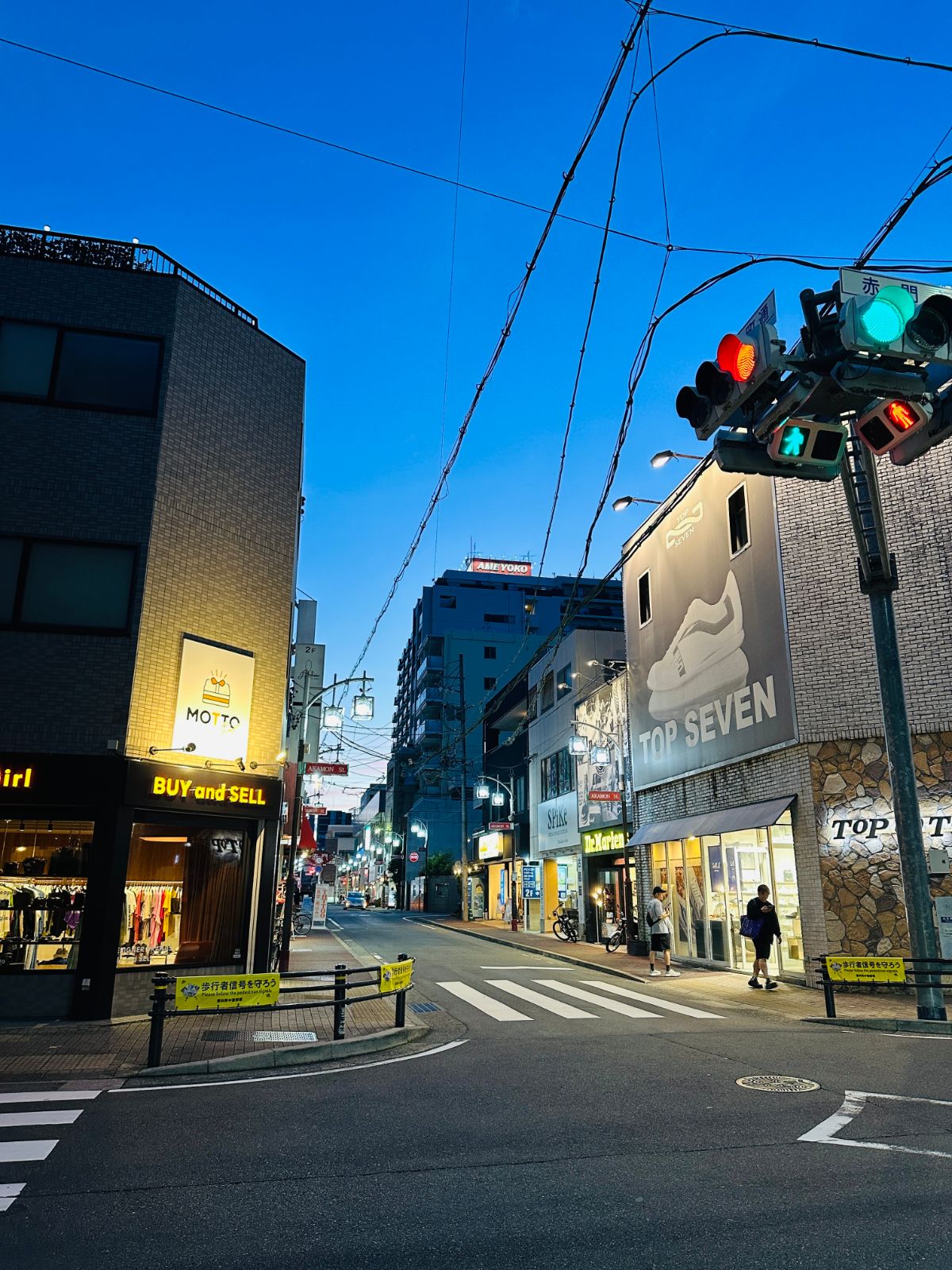 傍晚的日本街角，深藍色天空下交通號誌亮著綠燈，兩側是 TOP SEVEN 等店家招牌與交錯的電線