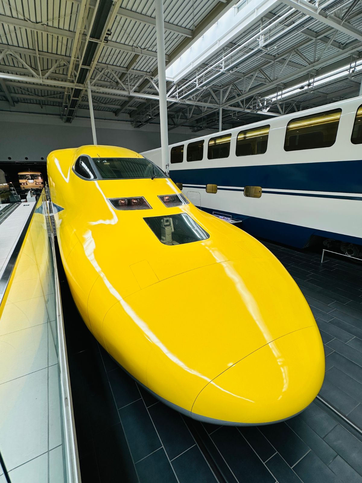 鮮黃色的新幹線試驗車車頭特寫，圓潤的流線外型在館內燈光下反光，旁邊停著藍白配色的舊型車廂