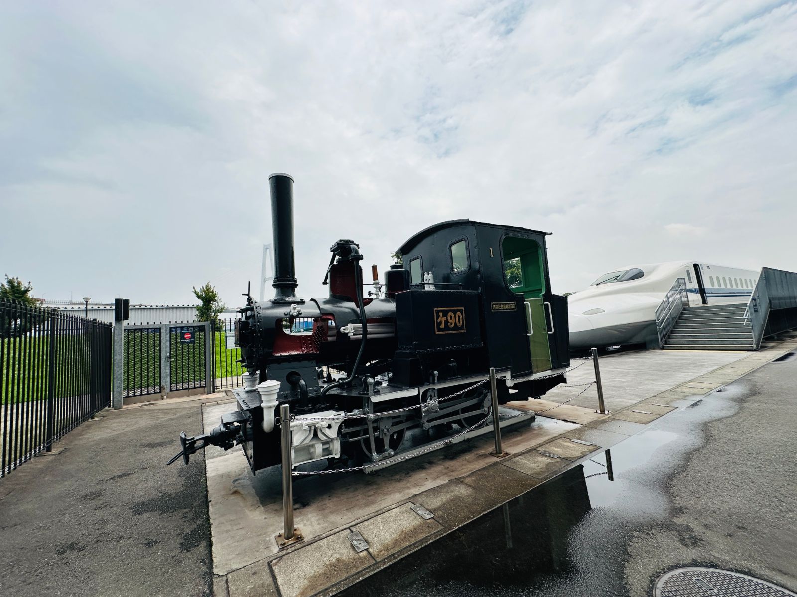 戶外展示的黑色蒸汽機車，車身標示「ケ90」，高聳的煙囪指向天空，後方停著白色新幹線車頭