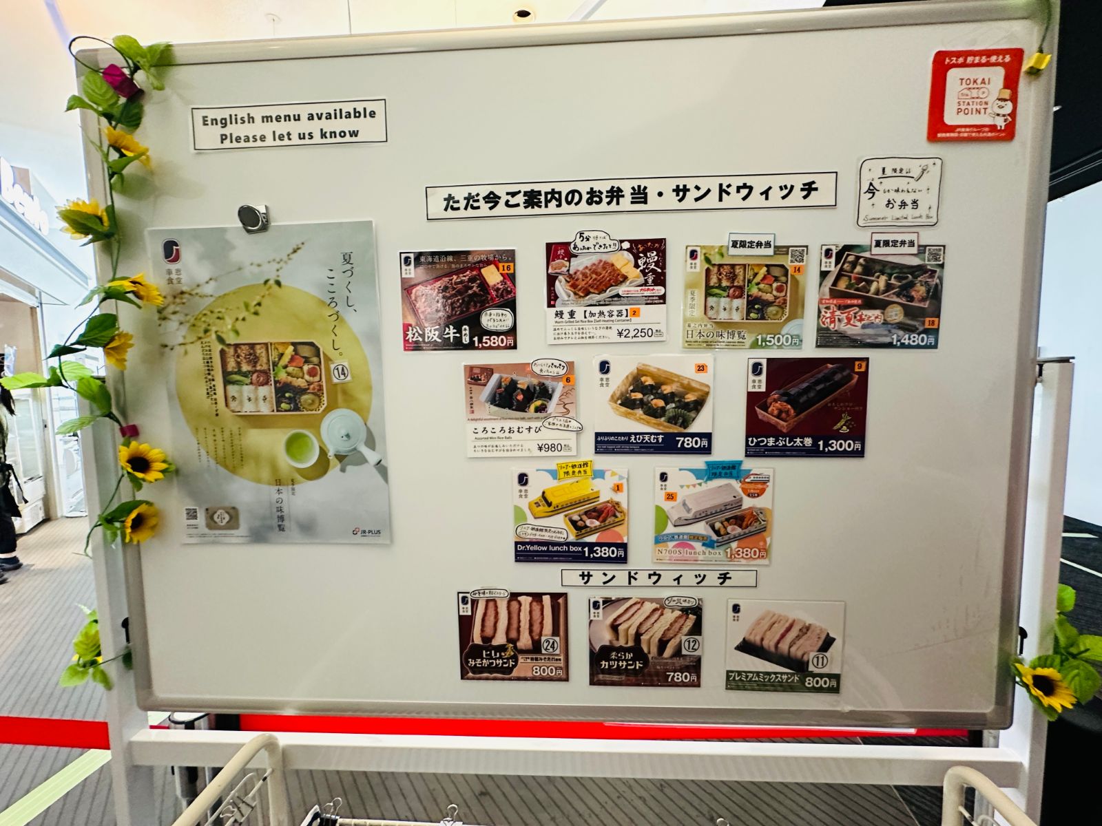 車站便當與三明治的販售看板，貼滿各式便當照片與價格，從 780 到 2250 日圓不等，兩側裝飾著向日葵