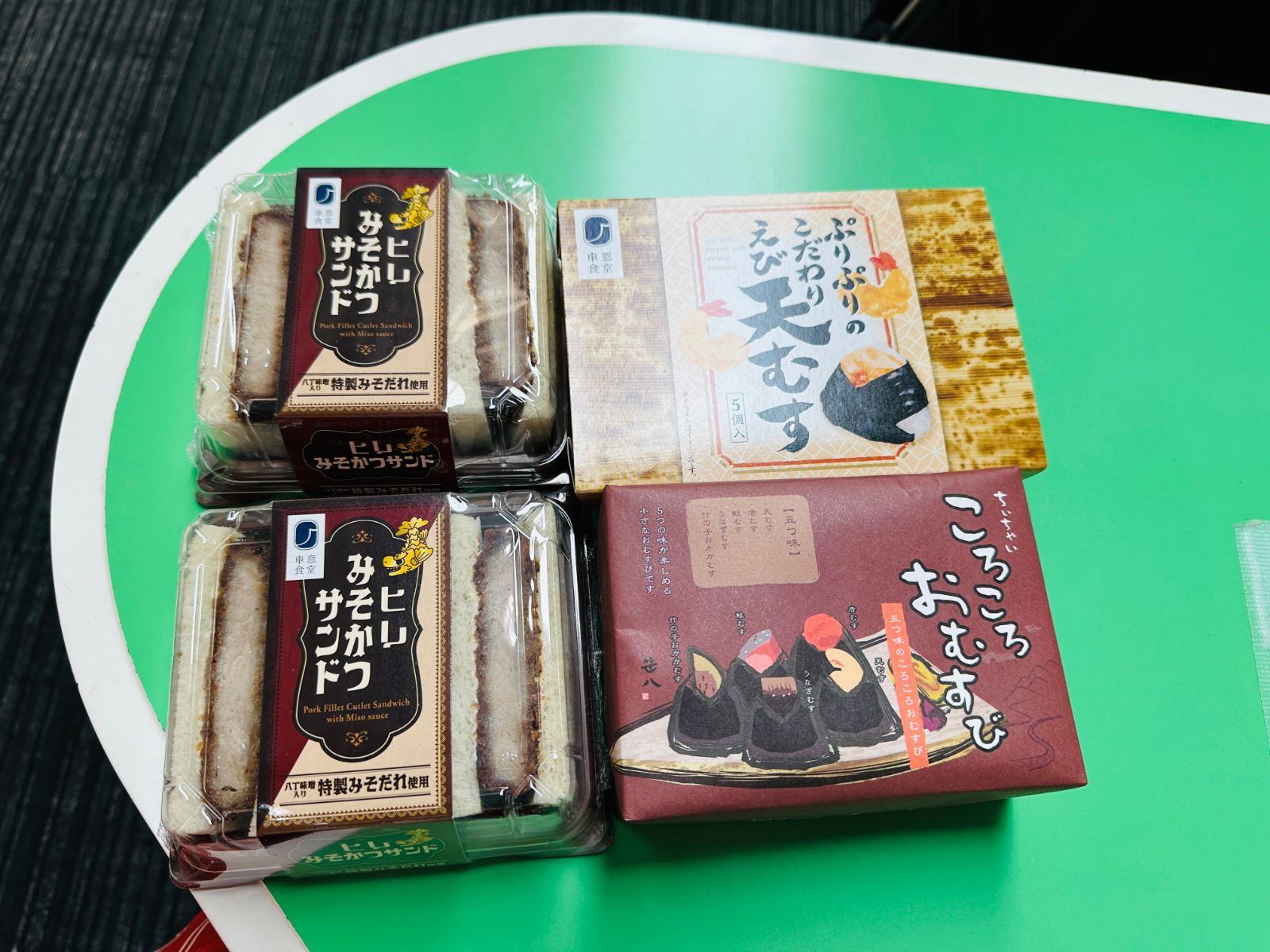 買回來的車站便當放在綠色桌面上，兩盒味噌豬排三明治、一盒蝦天婦羅飯糰與一盒綜合飯糰