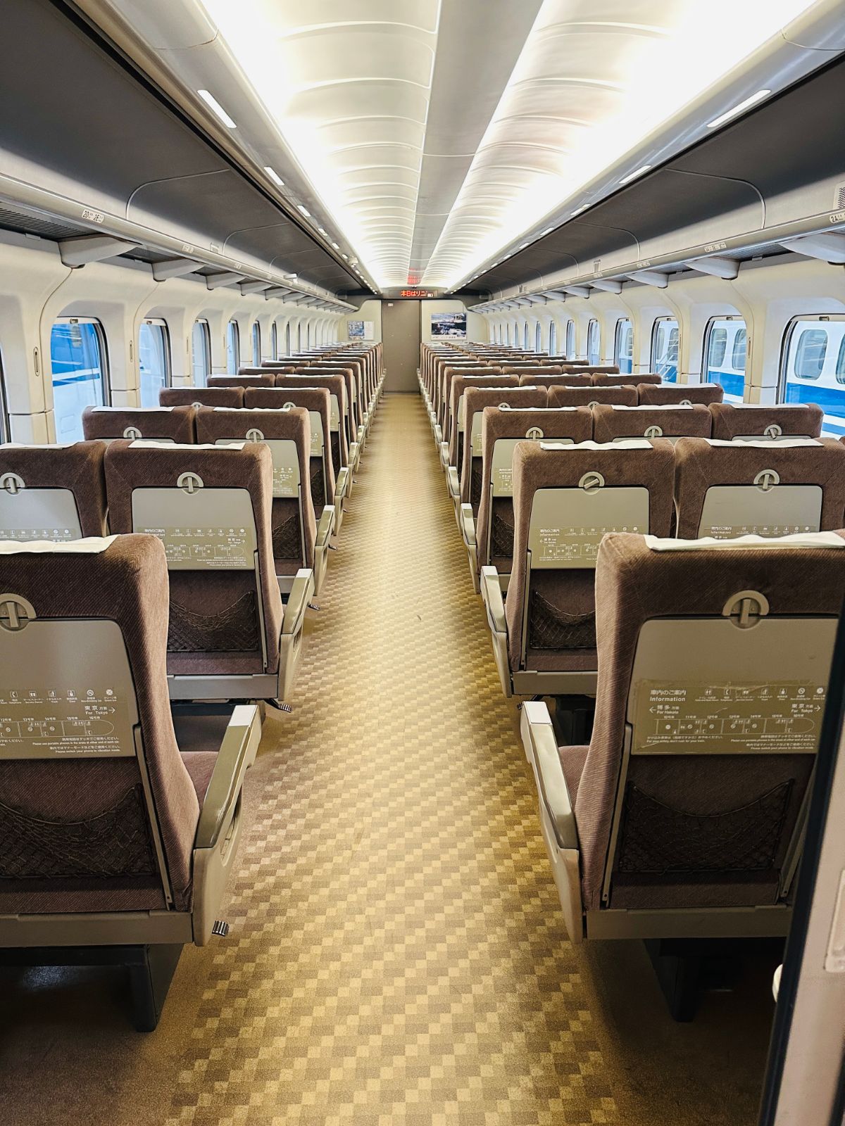 新幹線車廂內部，兩側三排咖啡色絨布座椅一路延伸到車尾，地板是米黃色格紋