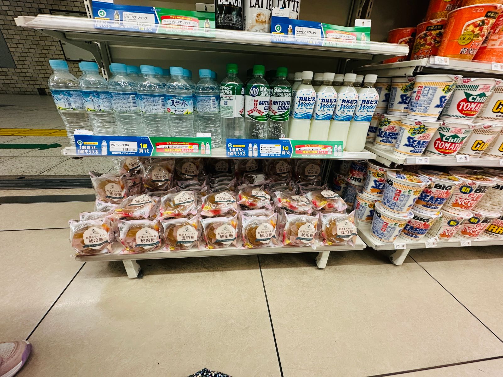 日本超商的貨架,上層排著礦泉水、可爾必思與各式瓶裝飲料,下層是包裝好的麵包與甜點,右側是整排杯麵